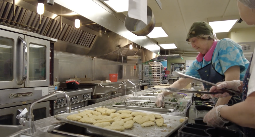 An assembely line of voulenteers in an industrial style kitching plating meals for Meals of Wheels of Syracuse.